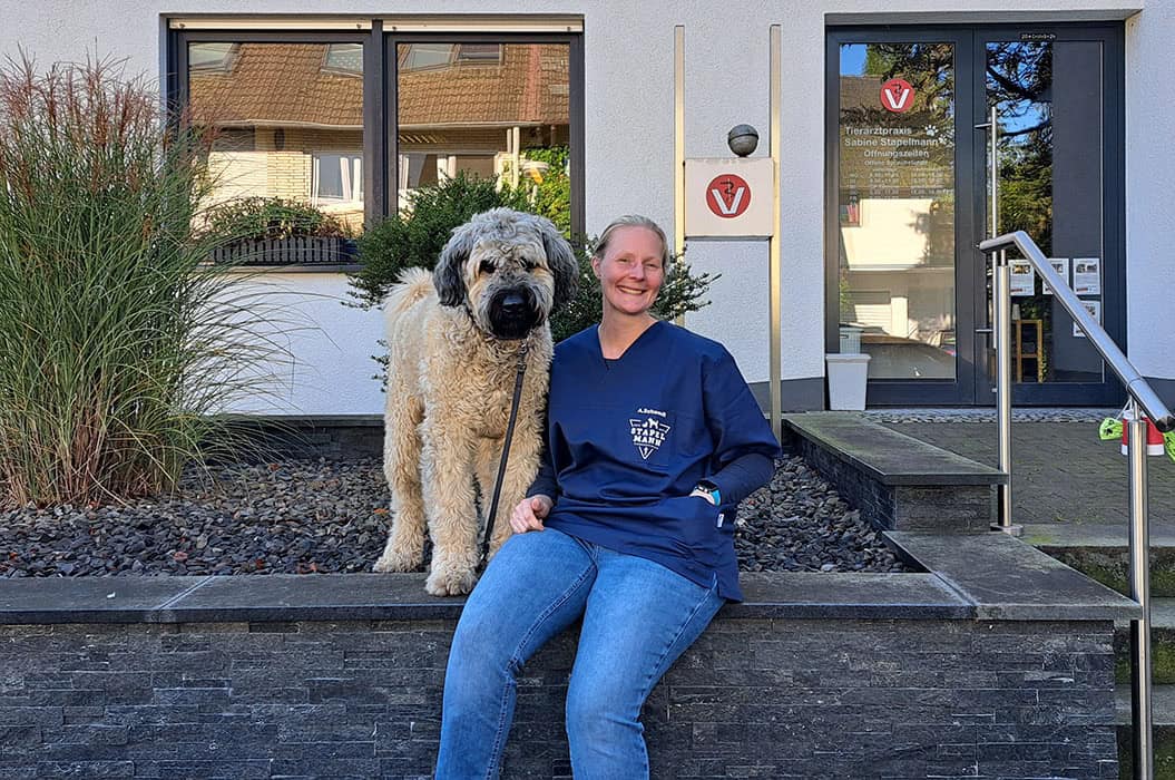 Eine lächelnde Person sitzt mit einem großen, zotteligen Hund auf einer Mauer vor einem Gebäude.